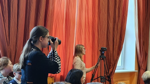 Viema skolniece fotografē, otra filmē skolas aktu zālē.