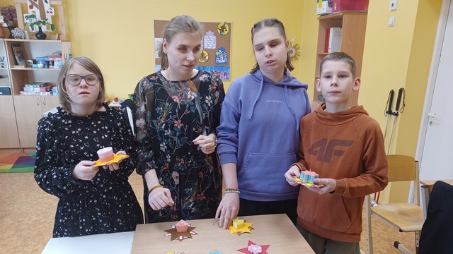 Trīs skolnieces un viens zēns stāv pie galda ar svecēm un arī rokās tur sveces.