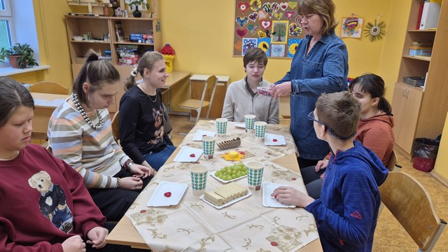 Skolotāja Inese pasniedz ēdienu skolēniem, kas sēž pie galda.