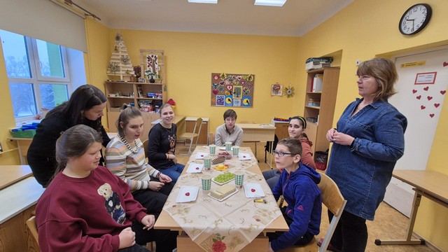 Skolēni pie galda, aiz viņiem - skolotājs, kas kaut ko stāsta.