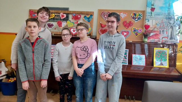 Piecu skolēnu grupa stāv pie klavierēm, uz kurām zīmējumi.