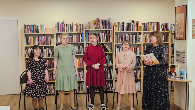 Četras meitenes dzied, skolotāja Inese blakus ar atvērtu grāmatu.