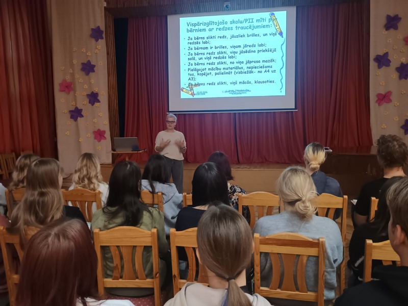 Direktore aktu zālē priekšā pie skatuves rāda prezentāciju un stāsta studentiem, kas sēž krēslos pret viņu.