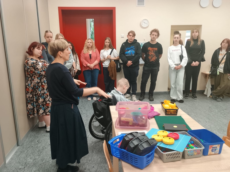 Skolotāja Airita klasē rāda speciālos palīglīdzekļus un viņai priekšā skolēns ratiņkrēslā.